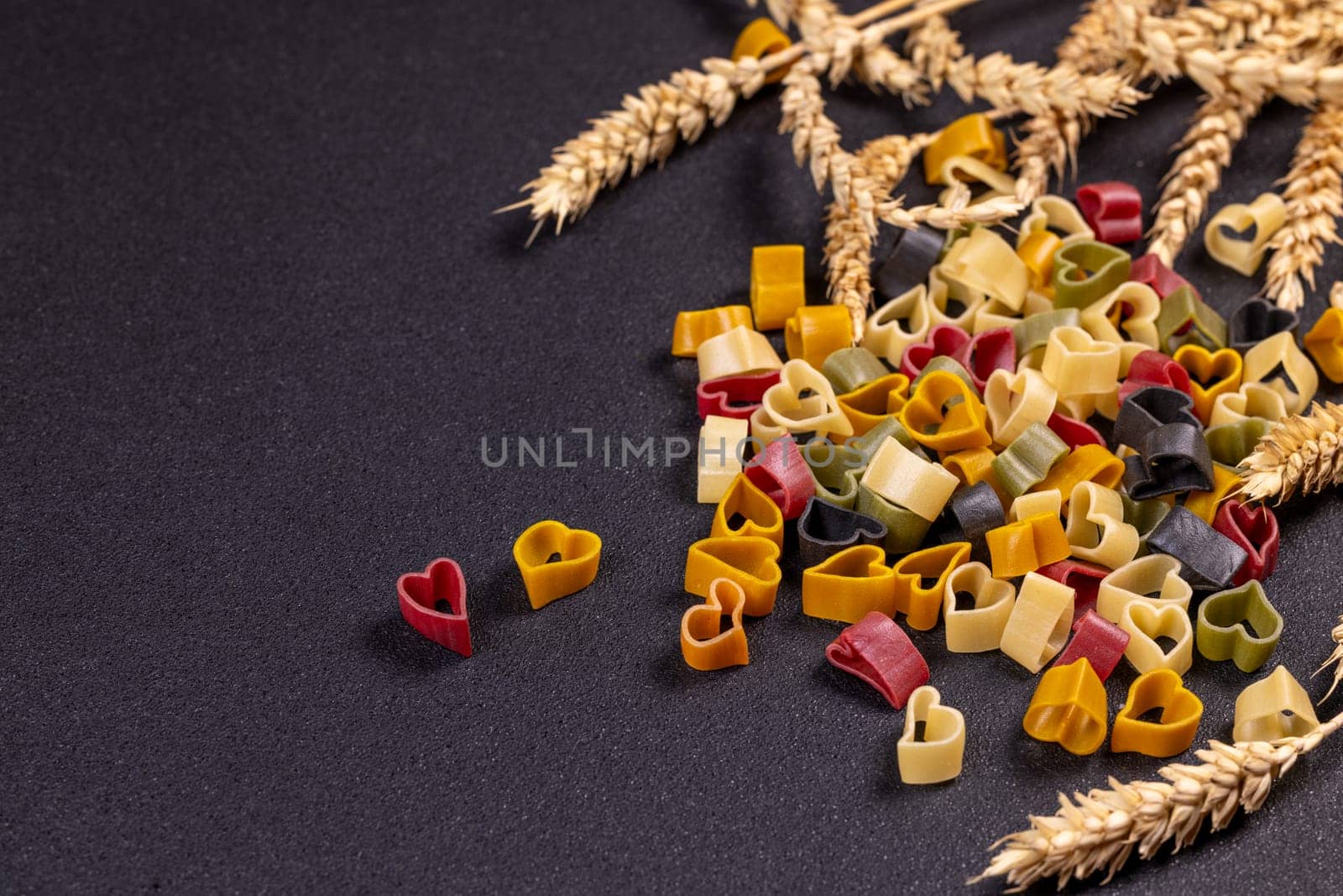 Small colorful heart shaped pasta is arranged on a black background with wheat, creating a romantic and culinary scene