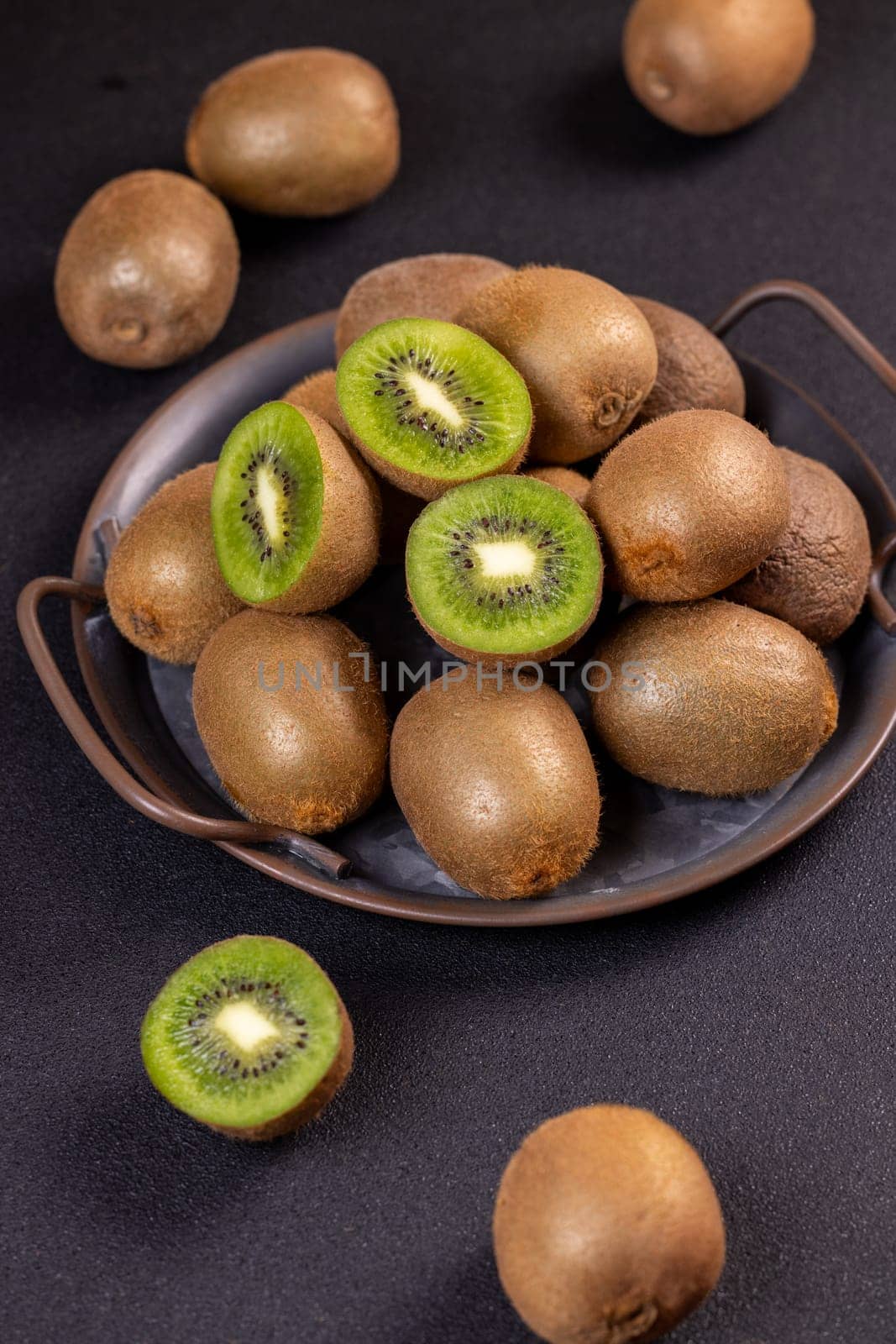 Ripe kiwi fruits are arranged on a metal tray and scattered on a dark surface, showcasing their fuzzy brown skin and juicy green flesh