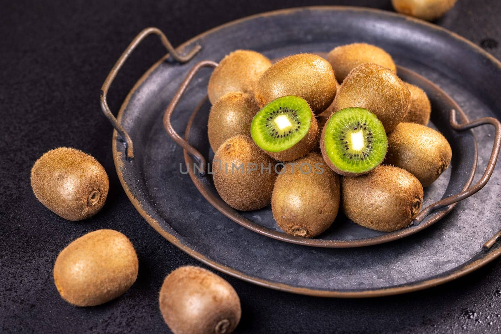 Ripe kiwi fruits are presented on a dark metal tray, showcasing their vibrant green flesh and fuzzy brown skin
