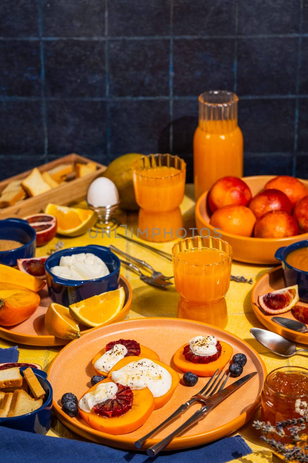 Slices of persimmon topped with ricotta, blood orange, and blueberries, arranged on a table with fresh fruit, juice, and toast