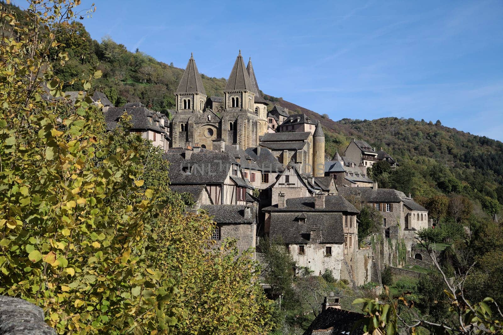 In Conques, France, medieval buildings stand tall among lush greenery under a clear blue sky.