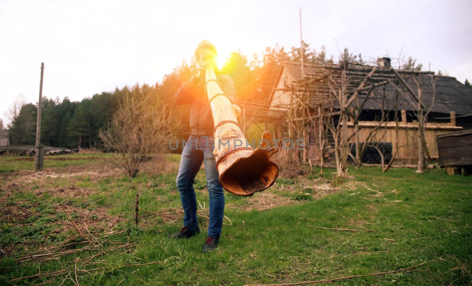 A man stands outdoors blowing into a large wooden horn, surrounded by a rural landscape and trees during a sunset.