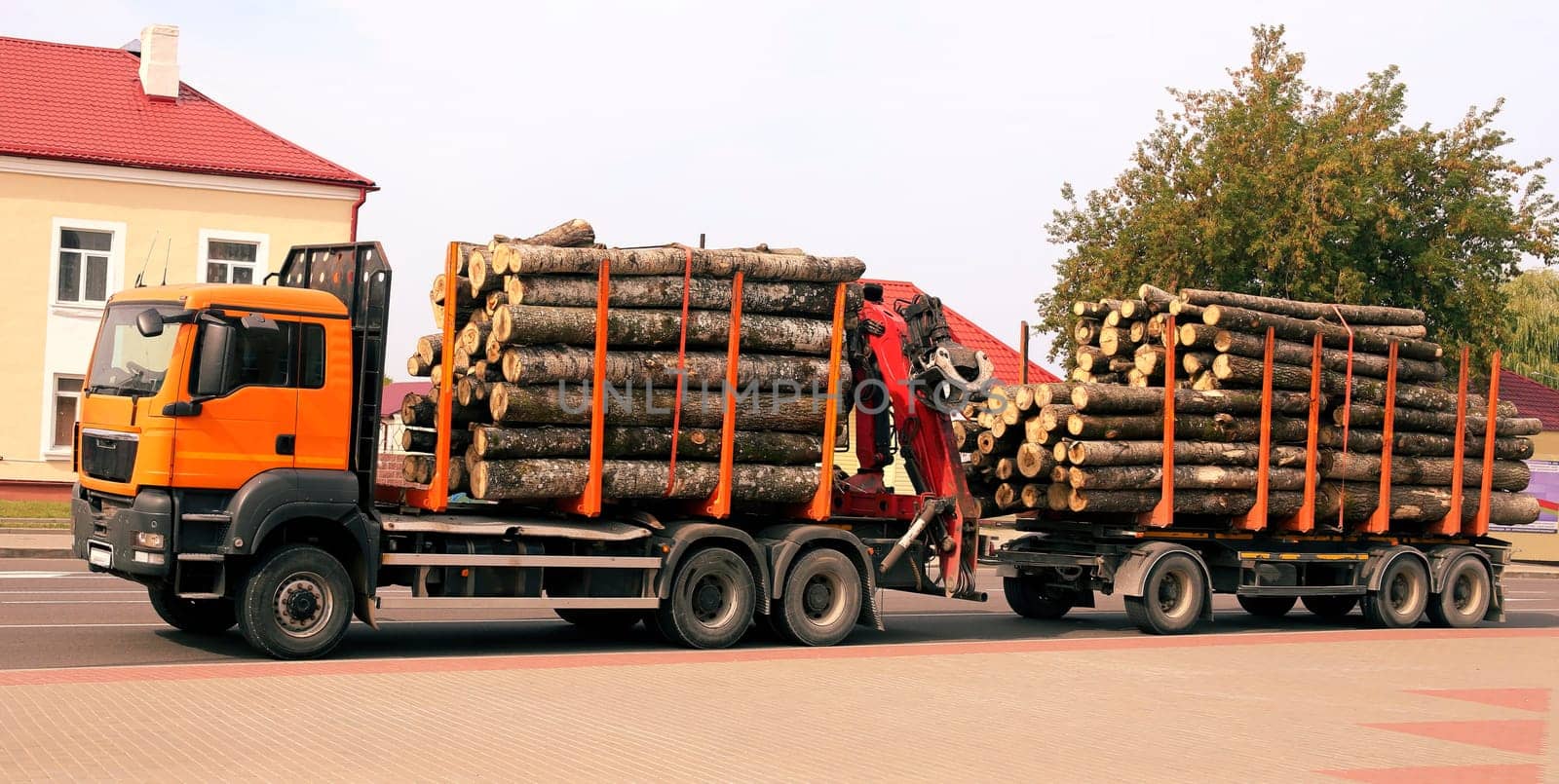 A flatbed truck loaded with stacked timber logs drives through a quiet rural setting on a sunny afternoon.