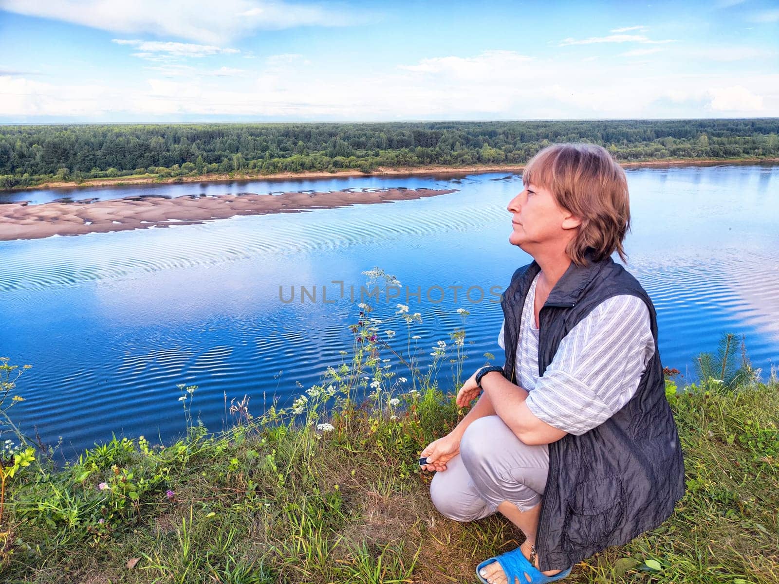 A middle-aged blonde woman kneels at a scenic riverside, capturing a selfie during her travel excursion in nature.