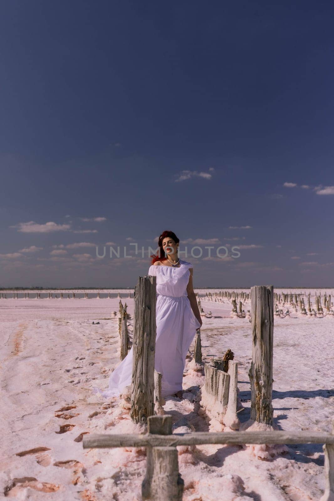 A woman in a white dress walks across a wooden structure on a salt flat. The sky is blue and there are clouds.