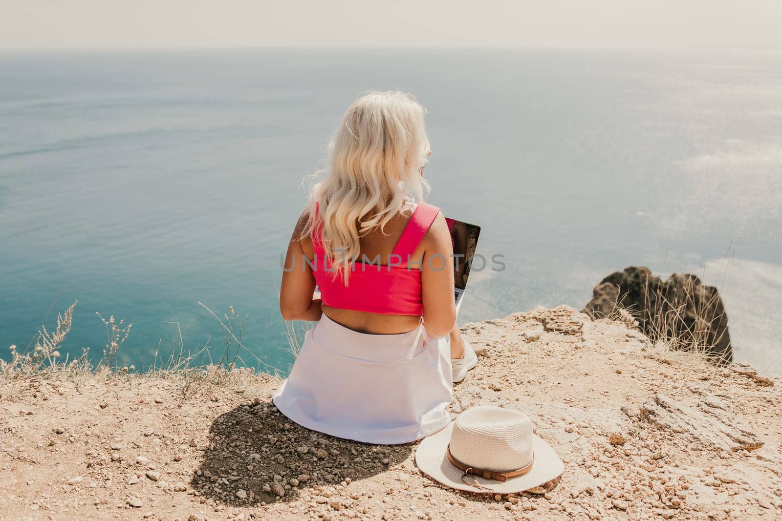 A woman is sitting on a rocky hillside with a laptop open in front of her. She is wearing a pink top and a straw hat. Concept of relaxation and leisure