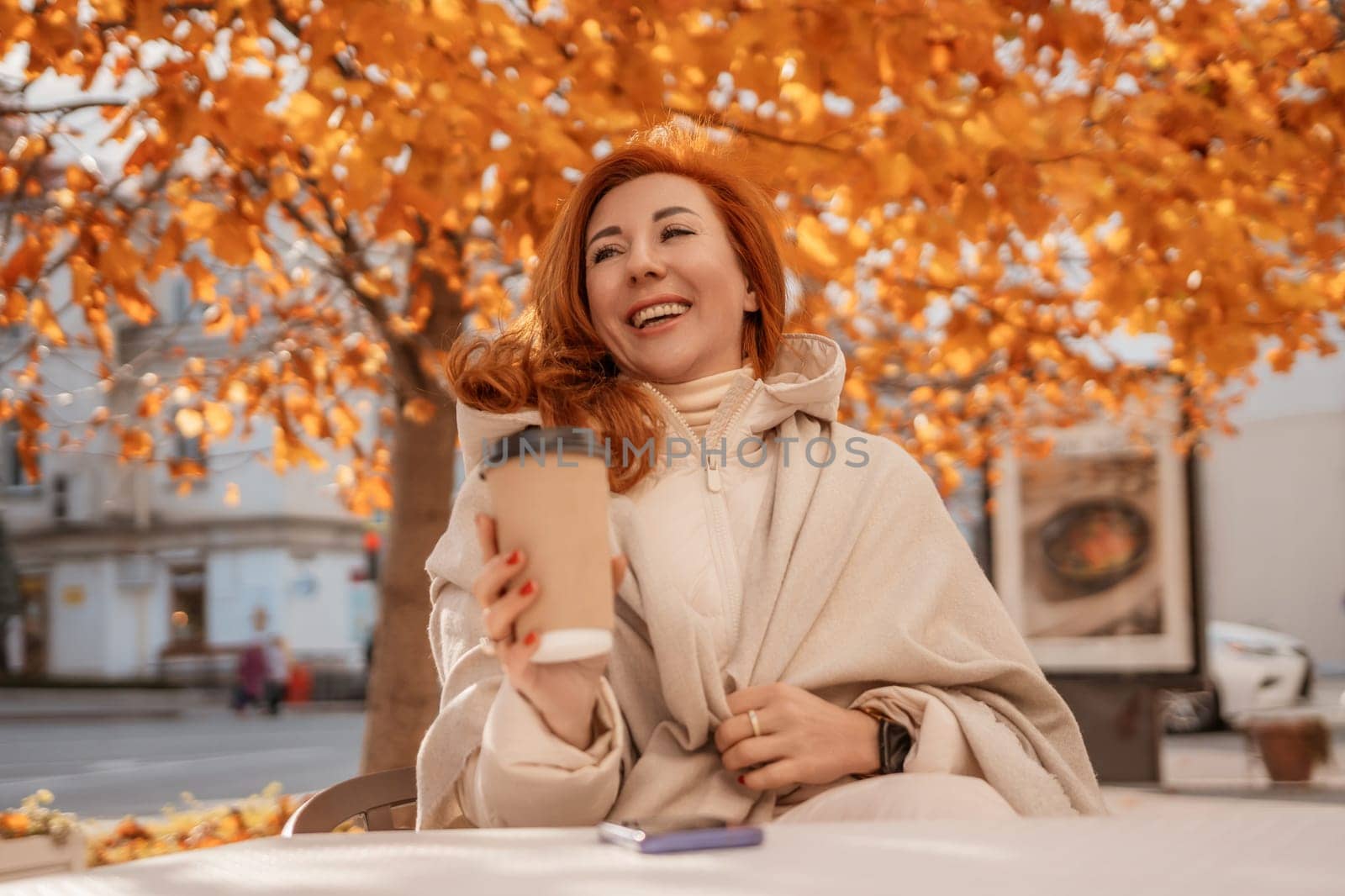Redhead woman smiles, wrapped in beige shawl, holding a coffee cup. Autumn leaves frame her under a tree. She sits at an outdoor table.