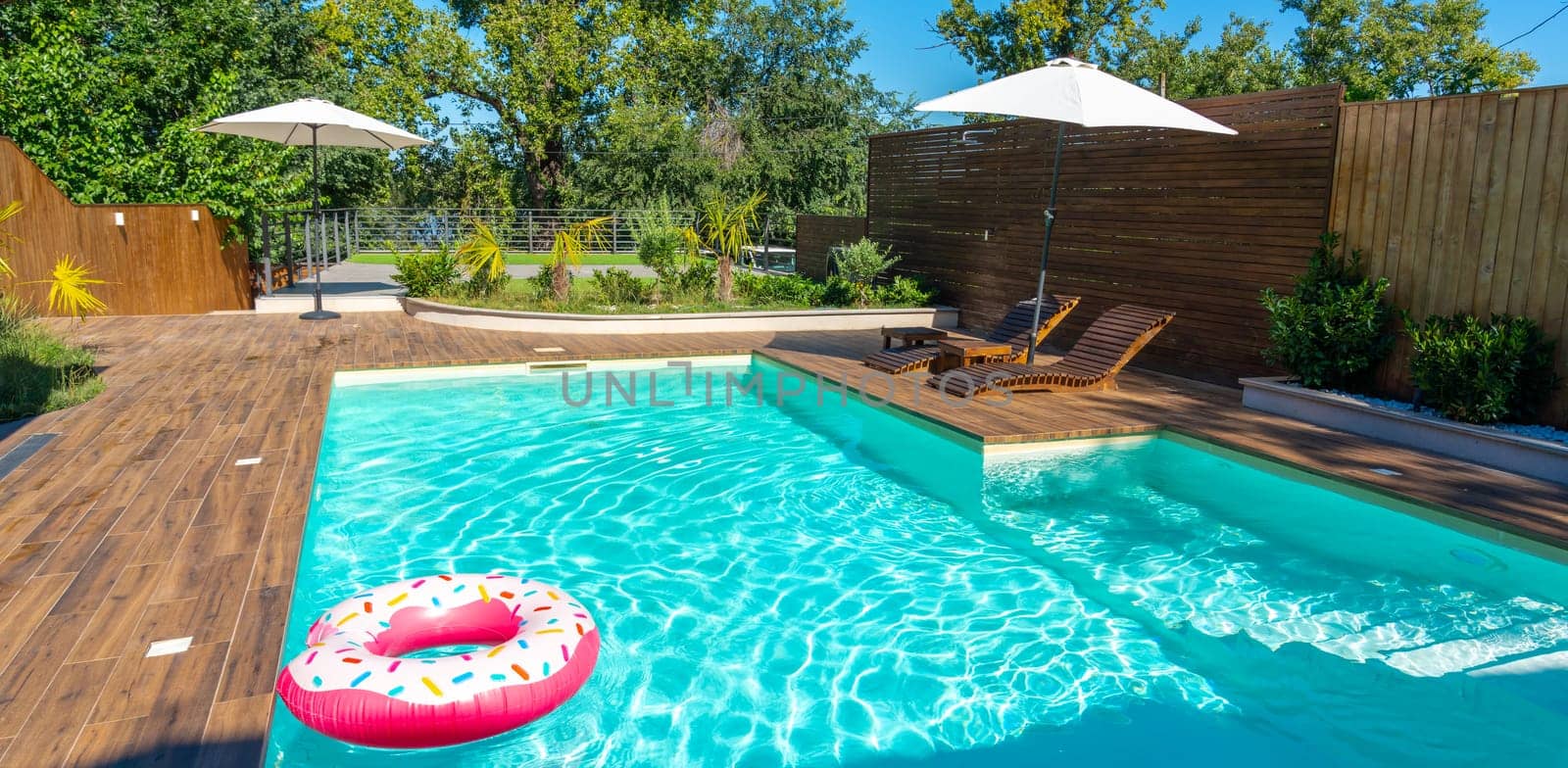 Modern backyard pool with turquoise water, wooden deck, umbrellas, lounge chairs, and pink inflatable ring
