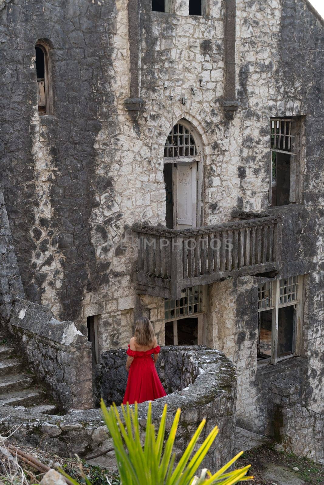 Woman in red dress stands near weathered stone castle ruins. Exterior features crumbling walls, decaying wooden balconies, and overgrown vegetation.