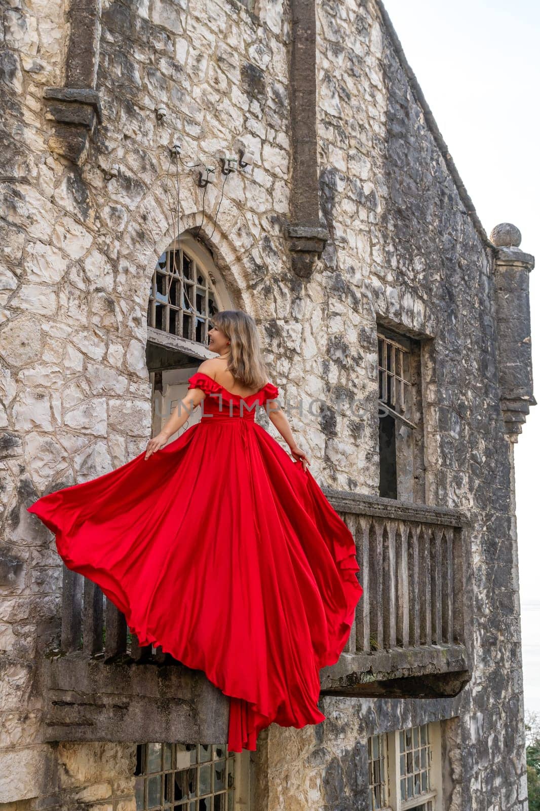 Woman in a flowing red gown stands on a weathered stone castle balcony, dress billowing in the wind.