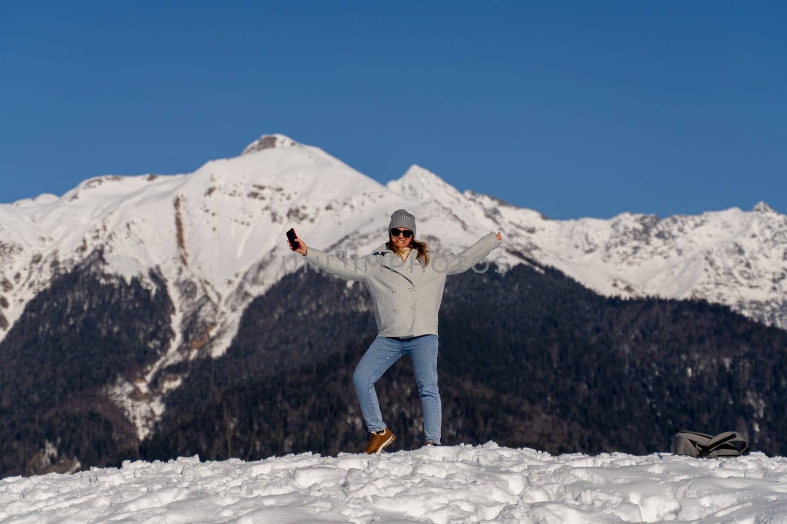 Woman in winter clothes joyfully poses with outstretched arms atop a snow-covered mountain peak against a backdrop of majestic snow-capped mountains under a clear blue sky.