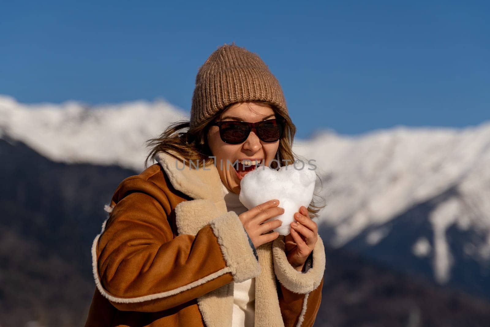Woman in brown coat and beanie playfully bites a heart-shaped snowball. Snowy mountains form a backdrop on a sunny day.
