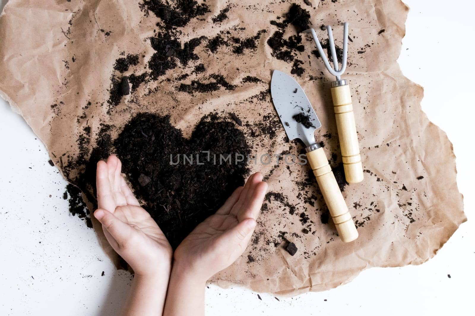 Child hands shaping soil heart with garden tools on brown paper.