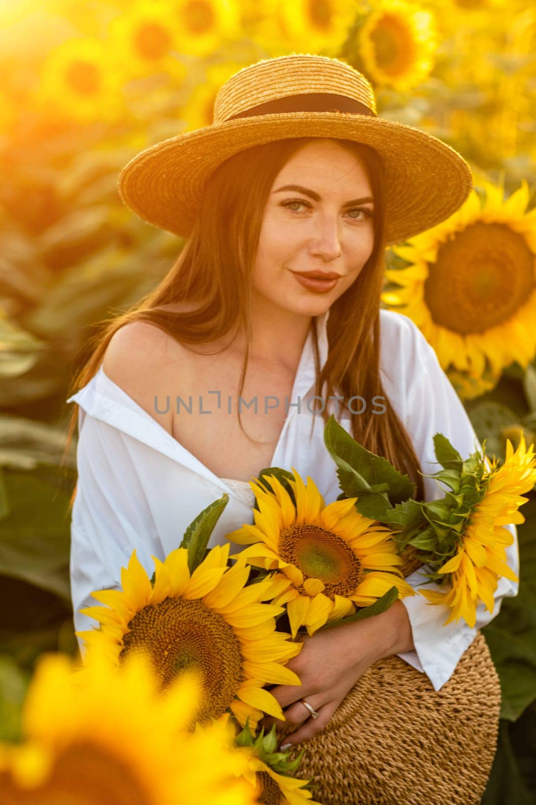 A girl in a hat on a beautiful field of sunflowers against the sky in the evening light of a summer sunset. Sunbeams through the flower field. Natural background