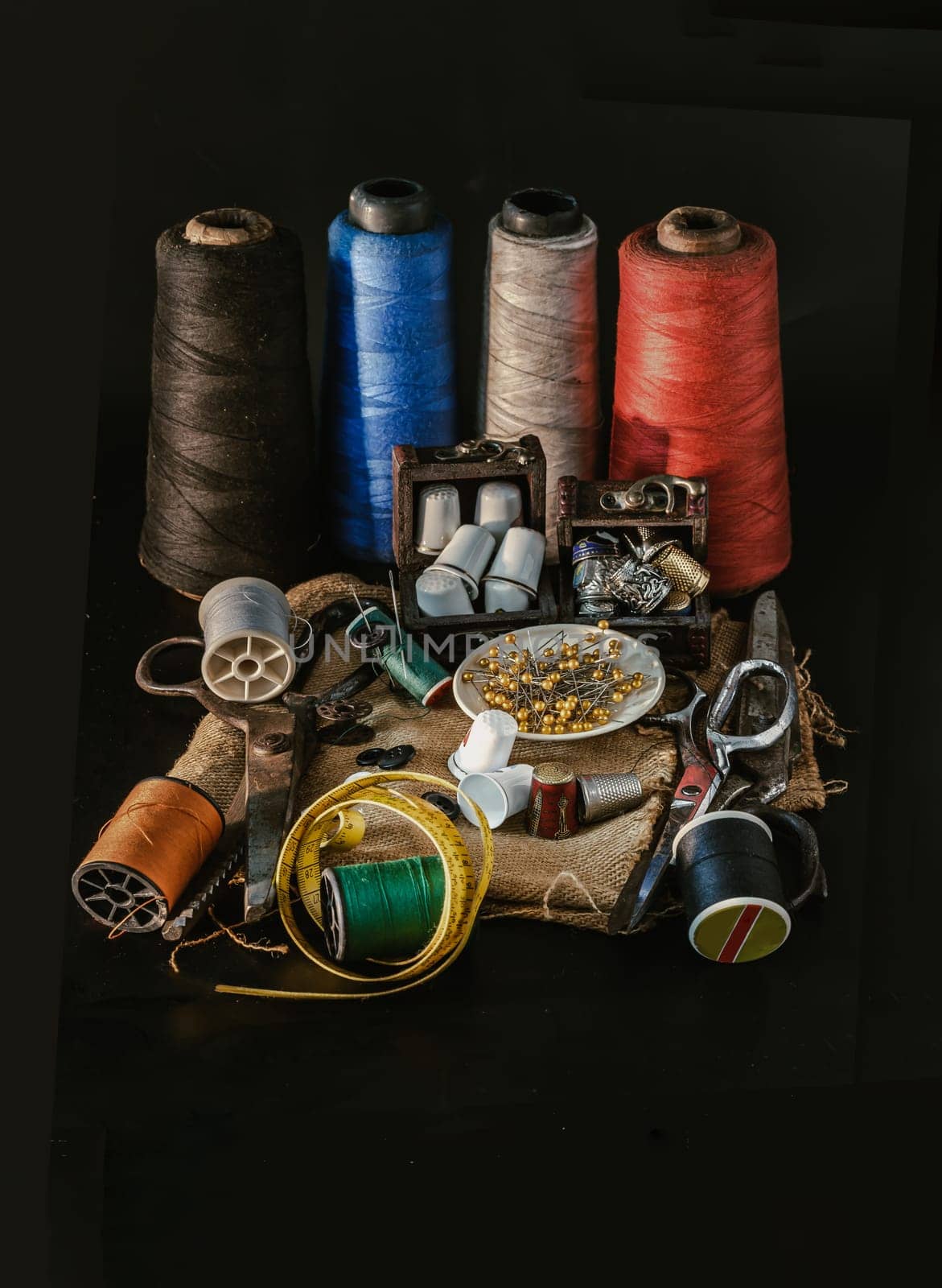 Thimbles in small wooden treasure chest, Spools of thread of various colour, Sewing needles stuck in a green miniature spools thread, Steel scissors and Measuring tape including pins in small round tray on Sackcloth with Black background. Sewing items set or Sewing notions (Sewing objects), use it as your Wallpaper, Poster and Copy space, Selective focus.
