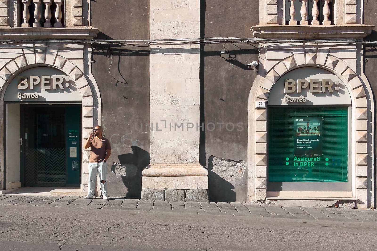 Catania. Italy - March 3, 2025: Striking BPER Bank facade with ornate stonework and a balcony, reflecting a clear blue sky. Features modern signage and security camera, ideal for finance, architecture