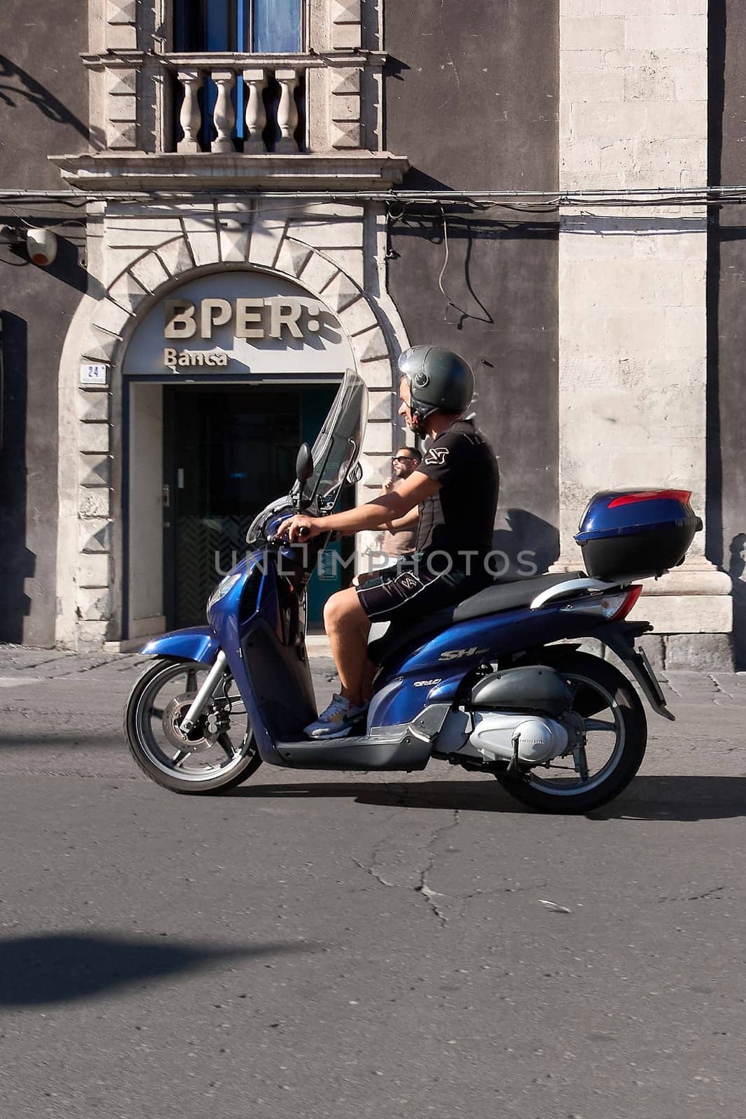 Catania. Italy - March 3, 2025: Energetic scooter rider in motion past the ornate BPER Bank facade with stone archway and balcony. Captures urban life, modern transport, and historic architecture.