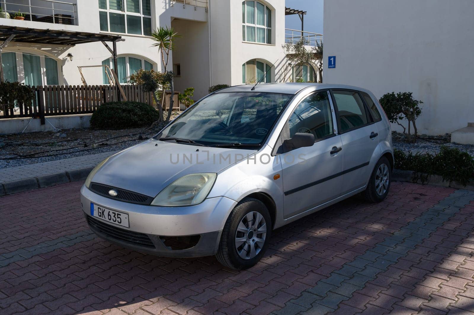 Gaziveren Cyprus -01.03.2025 A silver hatchback is parked on a paved surface, surrounded by sunny greenery and modern structures.