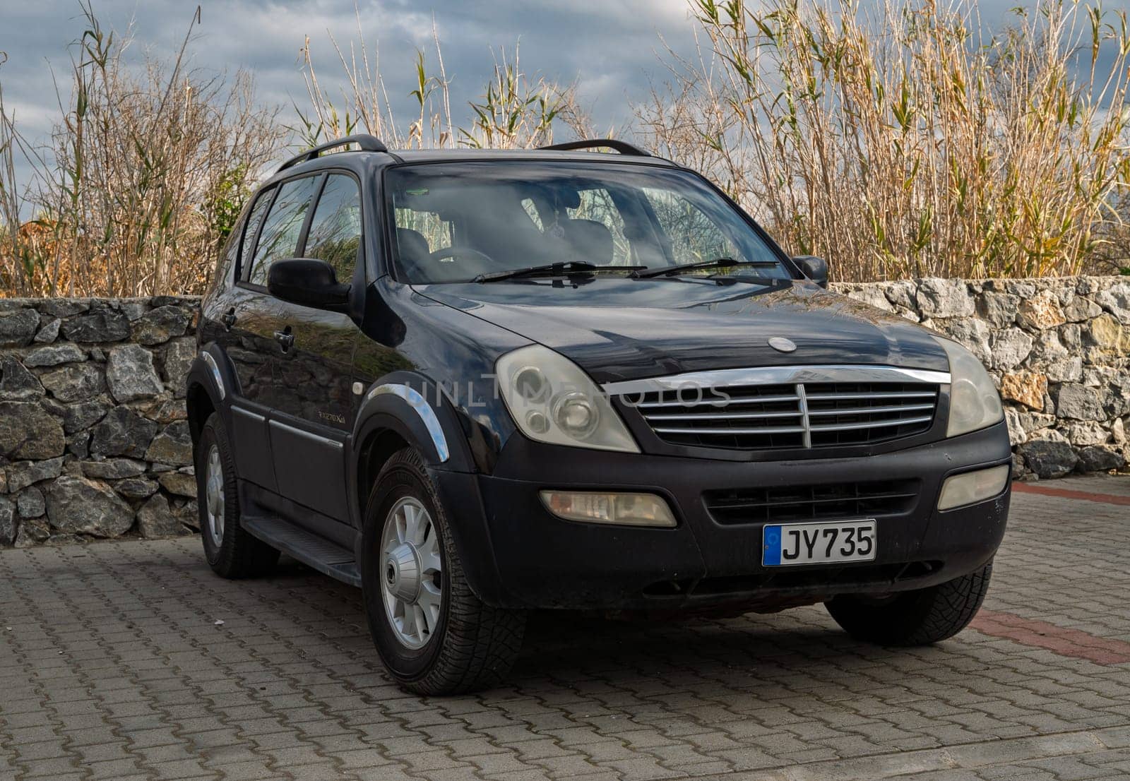 Gaziveren Cyprus -28.02.2025 A classic black SUV is parked on a paved area beside a rugged stone wall and tall greenery under cloudy skies.