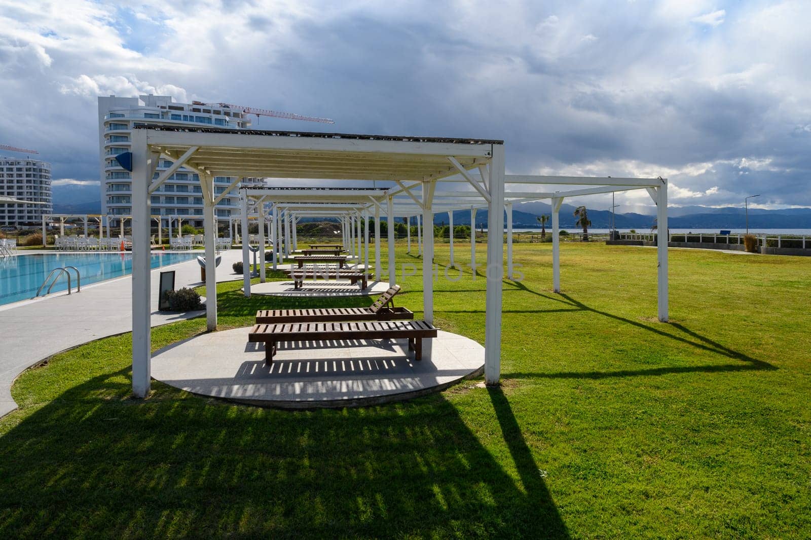 Shaded wooden benches line a serene pool area, framed by lush grass and vibrant clouds overhead, inviting relaxation.