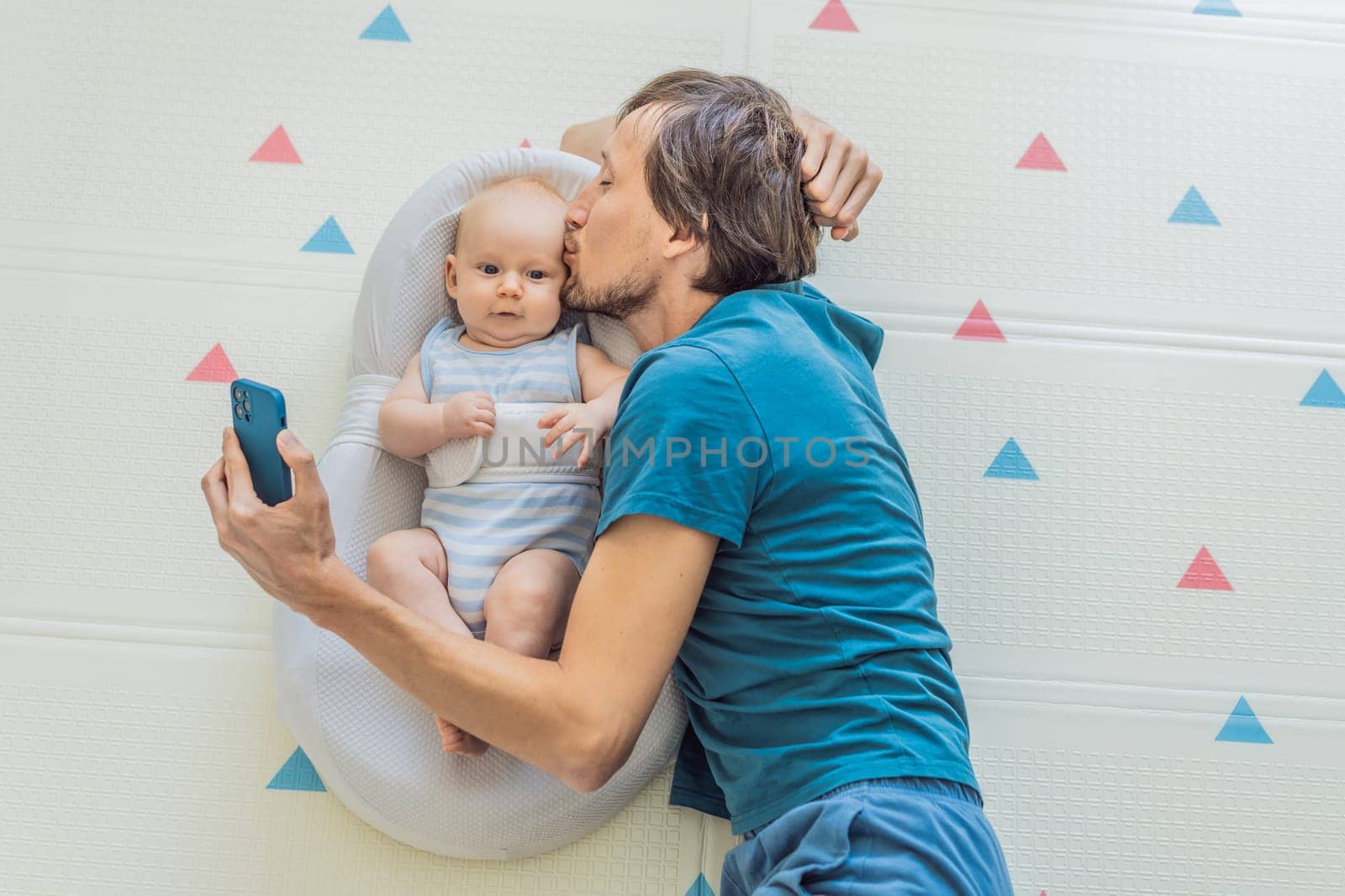 A 3-month-old baby lies safely in a cozy infant cocoon, secured with a special safety belt, while their father lies beside them during a video call. Modern parenting, bonding, and technology concept.