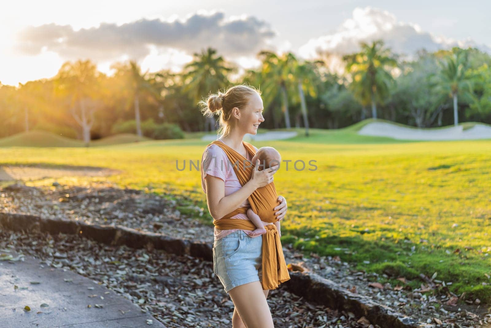 A loving mother walks through a peaceful park, carrying her baby securely in a sling. A heartwarming moment of maternal bonding, babywearing, and the joy of motherhood in nature.
