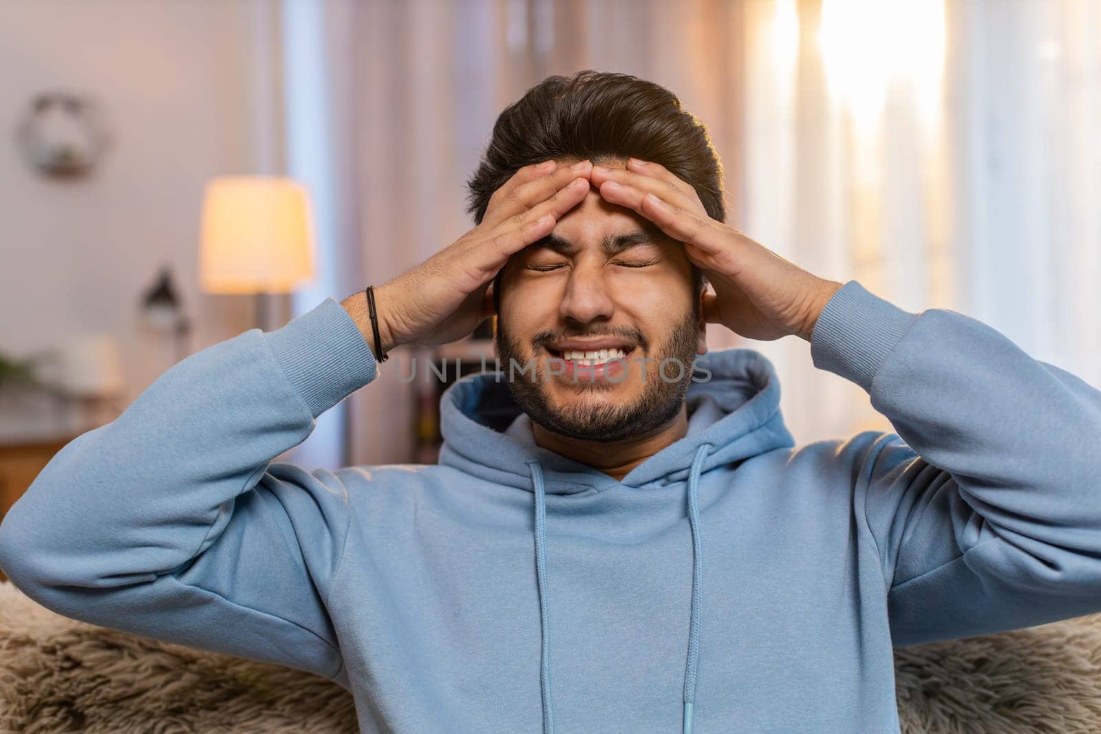 Sick Indian man rubbing temples to cure headache, suffering from tension, migraine, stress, and high blood pressure at home. Hispanic guy holds head, grimacing in pain, experiencing severe discomfort.