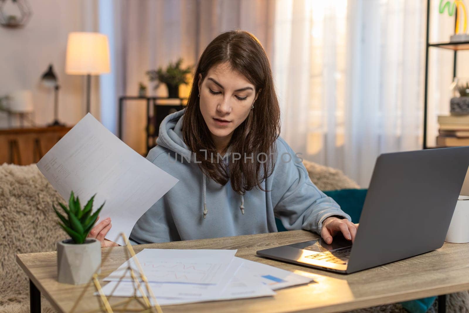 Young Caucasian woman sitting at table with laptop working on business plan looking focused typing text with documents spread around. Confident girl freelancer working from home in apartment room.