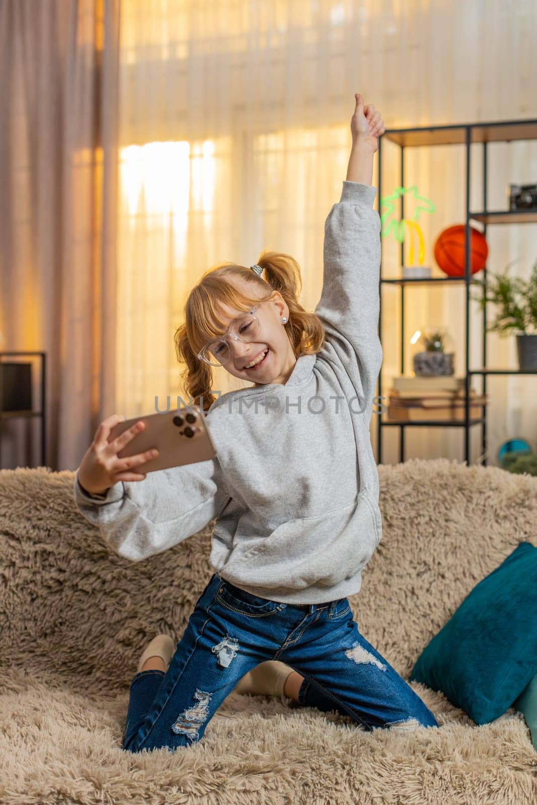 Young teenager girl using smartphone at home, giggling while playing passing a tough online game level with excitement. Child kid on sofa waving phone in joy, celebrate win victory cheerful triumph