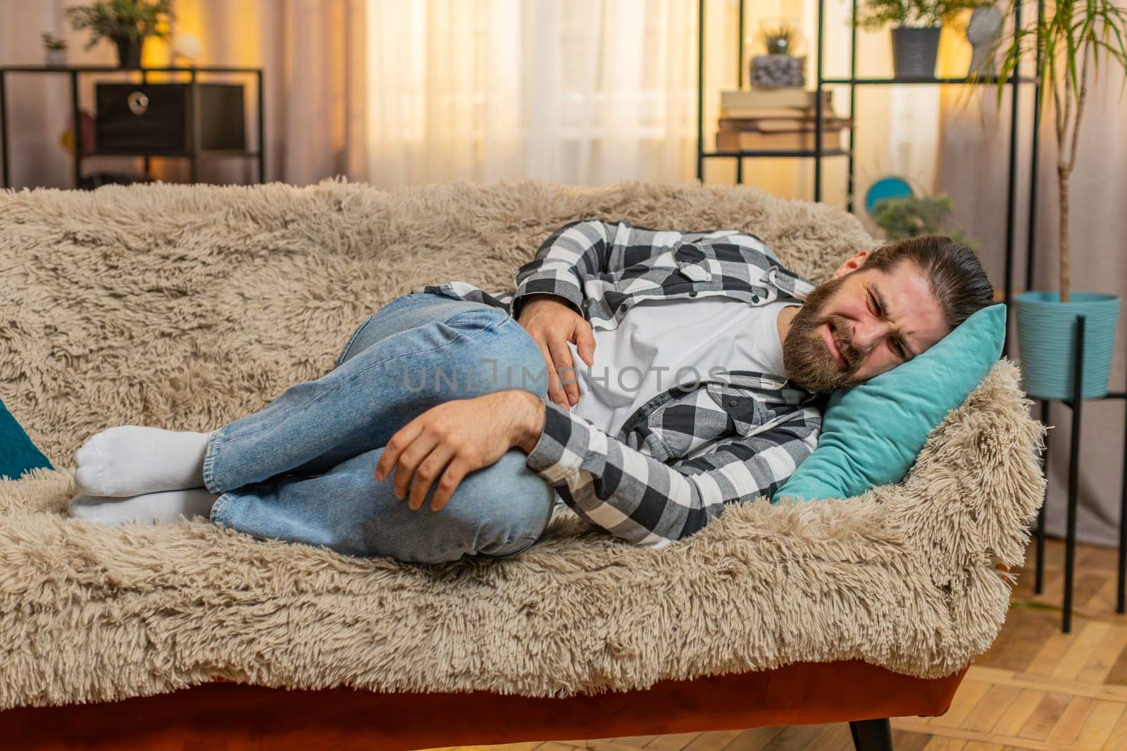 Young ill sick man lying at home with palm on abdomen, quietly moaning from stomach trouble or overeating. Caucasian guy on sofa curls slightly, enduring pain from potential gastritis or diarrhea.