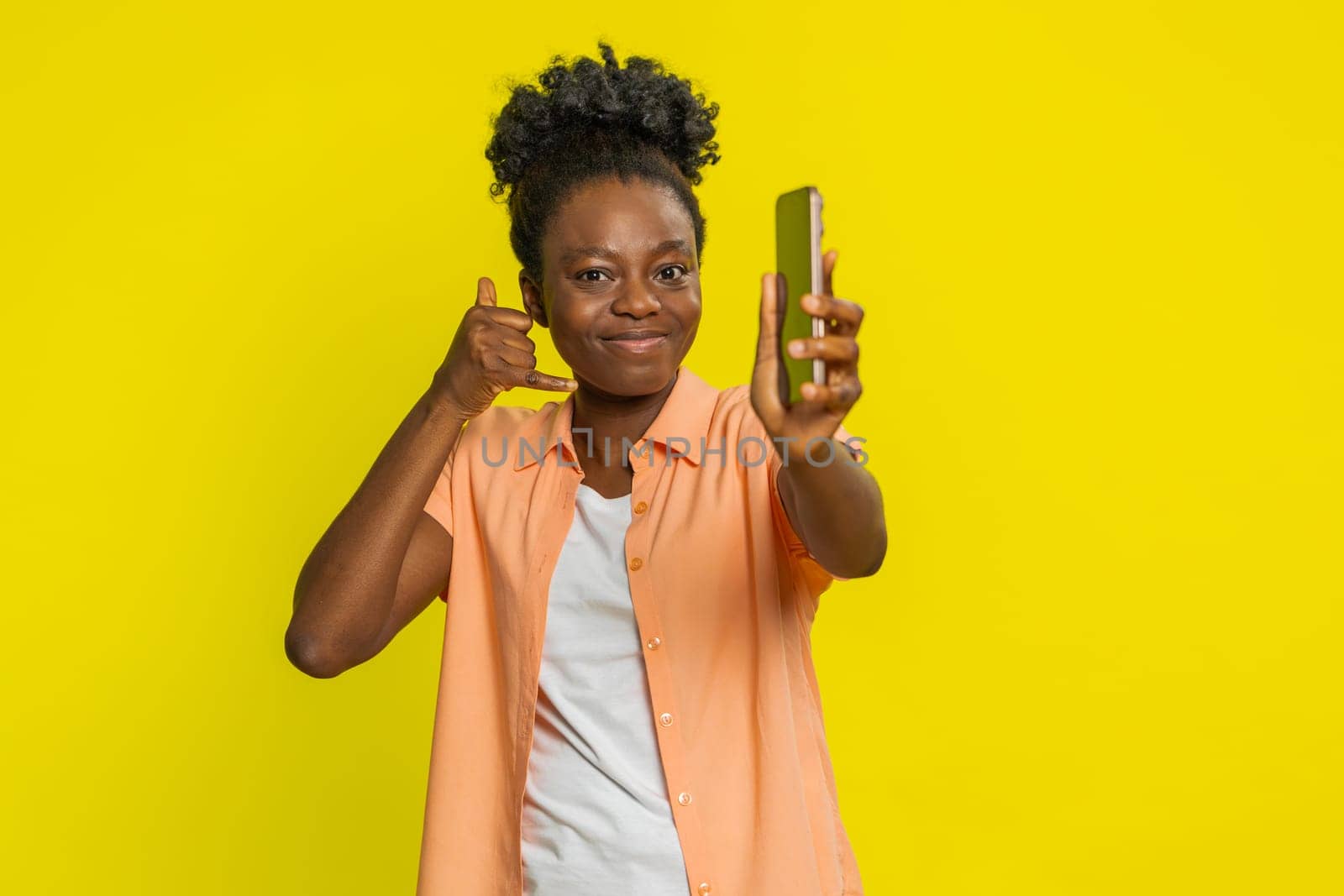 African American woman holds smartphone and makes call me gesture, signaling a desire for connection. Black girl on yellow background radiates confidence and highlights direct invitation to contact