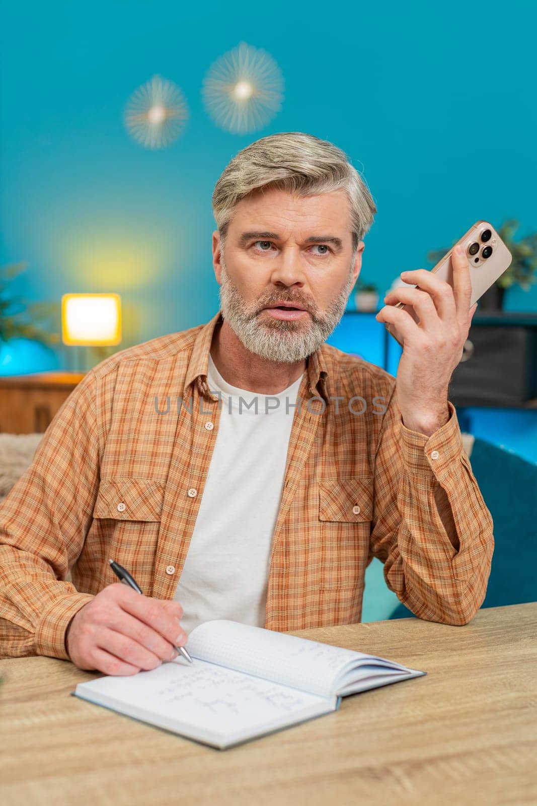 Mature man talking on mobile phone loudspeaker, making business discussion and writing notes in notebook. Middle-aged guy sitting at home table focusing on solutions, feeling determined. Vertical