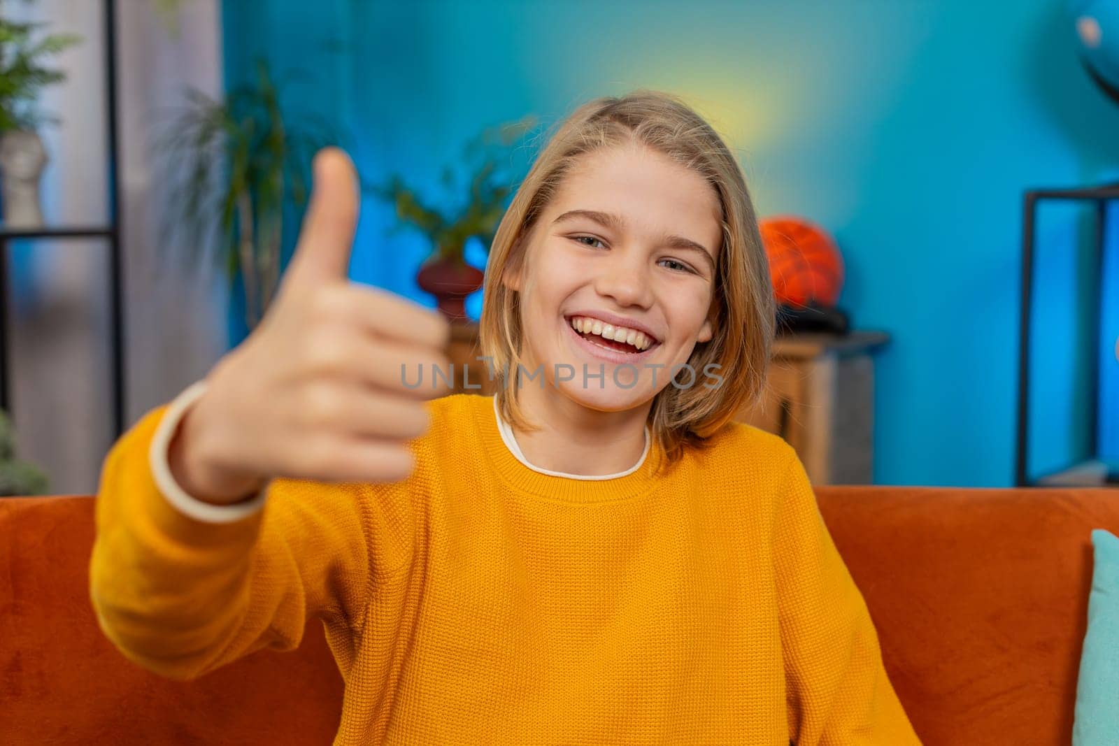 Like. Caucasian teenager boy gives thumbs up to camera while smiling at home. Sits comfortably on sofa, appearing satisfied with the good positive result, displaying feelings of approval, contentment