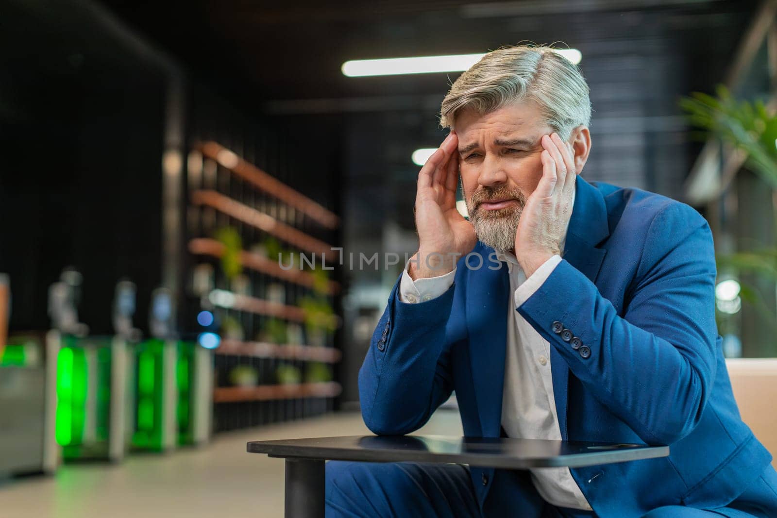Tired ill middle-aged businessman suffering from headache problem tension and migraine, stress in modern office lobby. Handsome freelancer businessperson man guy sitting in hotel hall. Overworking.