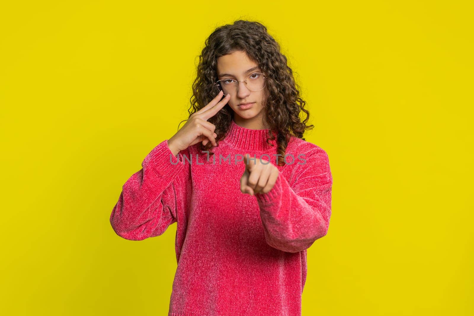 I am watching you. Confident attentive Caucasian young teenager girl pointing at eyes and camera, show I am watching you gesture, spying on someone. Strict teen child isolated on yellow background