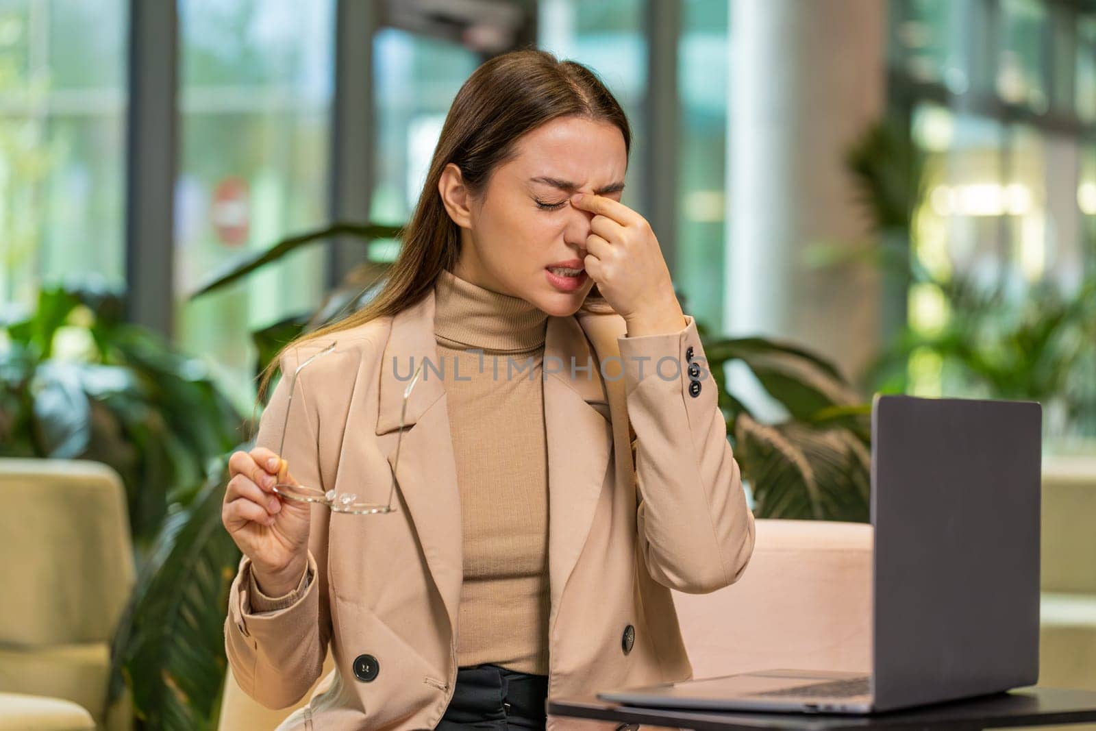 Overworked young Caucasian businesswoman suffering from eye strain and headache working on laptop sitting in office lobby. Tired female manager takes off glasses and massages, rubs bridge of nose.