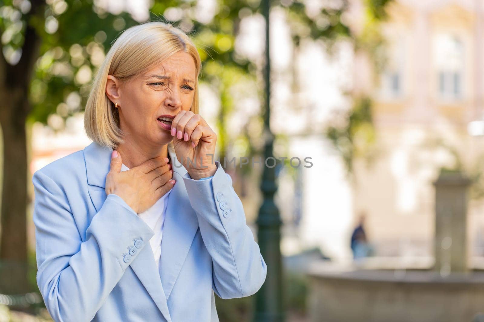 Sick mature businesswoman in formal suit suffering from sore throat while standing on city street. Upset Caucasian female manager with a cold or flu coughing and feels bad. She has a cold or virus.