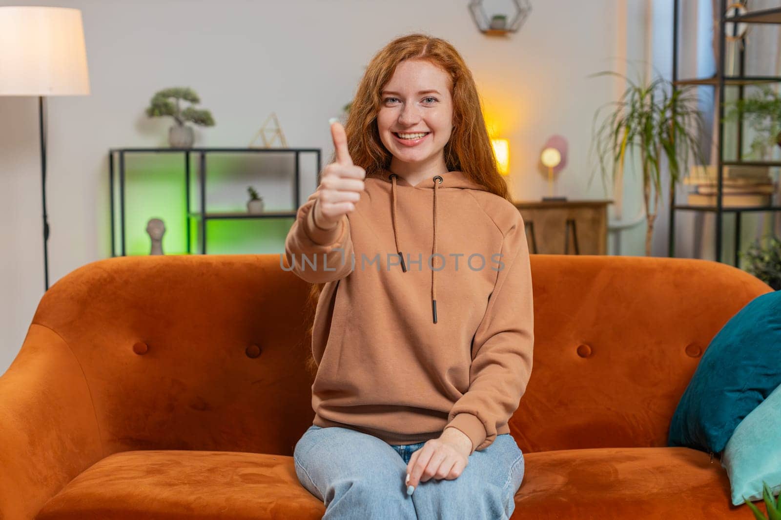 Portrait of excited smiling Caucasian woman looking approvingly at camera showing double thumbs up, like sign, good news, positive feedback. Happy redhead girl sitting on sofa in living room at home.