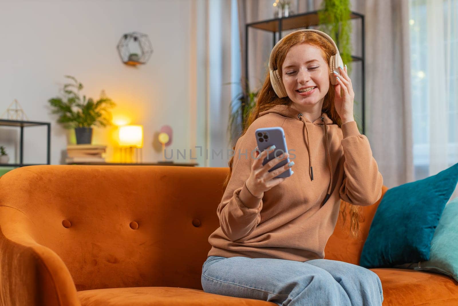 Happy overjoyed young redhead woman in wireless headphones relaxing sitting on couch at home apartment choosing, listening favorite energetic disco dancing music. People weekend leisure activities.