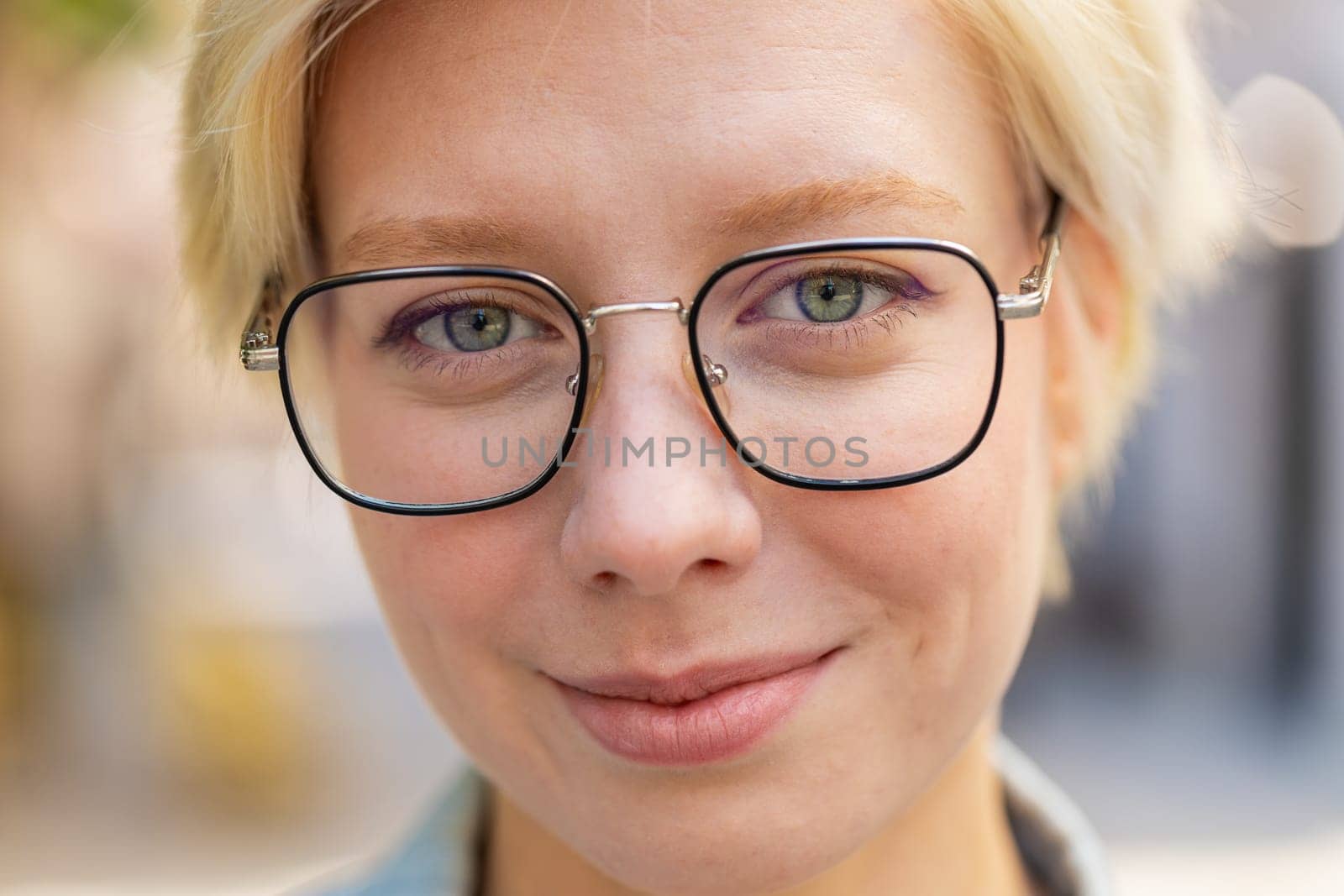 Close-up portrait of happy young woman face smiling friendly looking at camera resting relaxation feel satisfied good news outdoors. Pretty adult lady in glasses with short blond hair on city street.