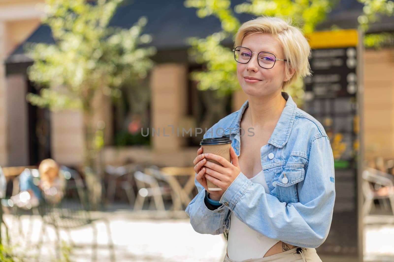 Happy Caucasian young blonde woman enjoying morning coffee hot drink and smiling outdoors. Relaxing, taking a break. Girl walking on urban city center street, drinking coffee to go. Town lifestyles.