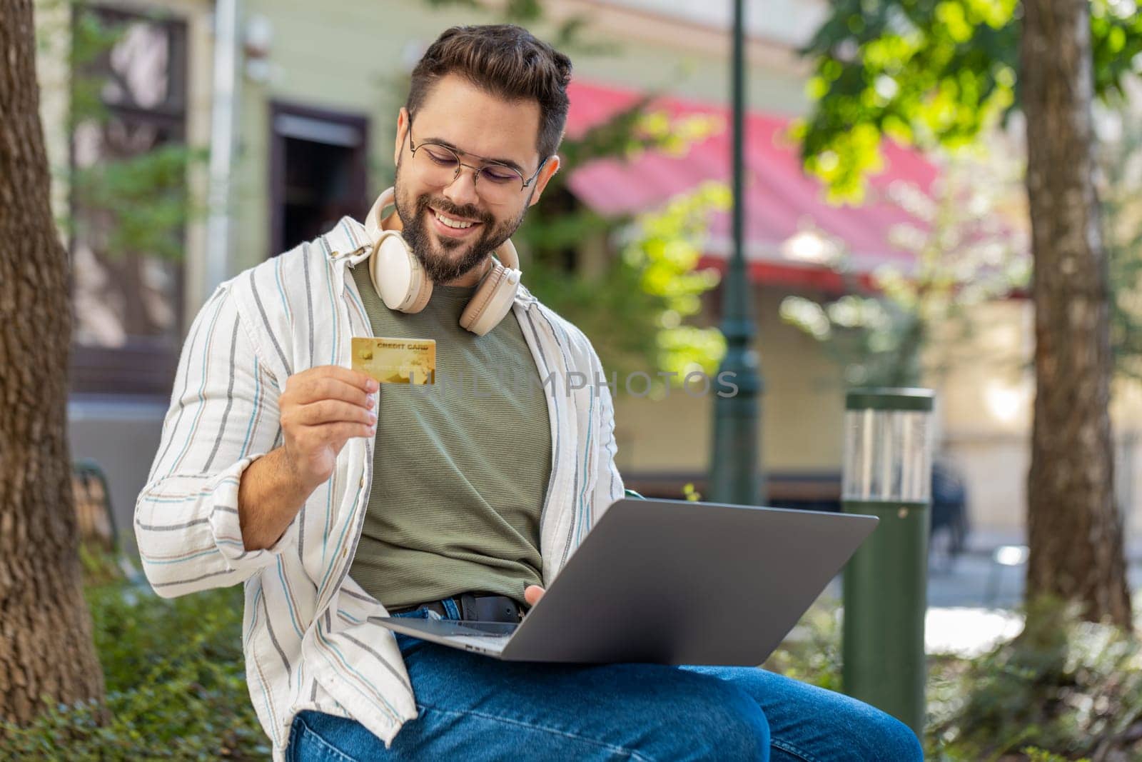 Caucasian young man using credit bank plastic card and laptop computer while transferring money purchases online shopping order food delivery on internet outdoors. Guy using netbook on city street.