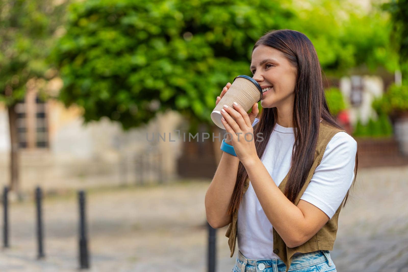 Happy Caucasian young pretty woman enjoying morning coffee hot drink and smiling outdoors. Relaxing, taking a break. Girl walking on urban city center street, drinking coffee to go. Town lifestyles.