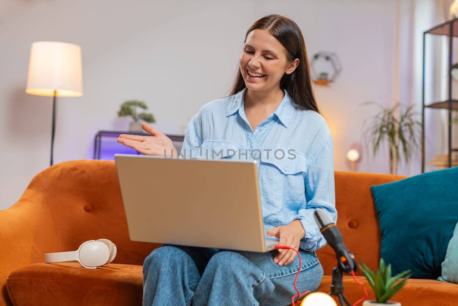 Happy blogger woman recording podcast at desk, giving workshop, webinar. Young Caucasian girl speaking at microphone and laptop webcam, making video call, discussing at home living room sits on couch