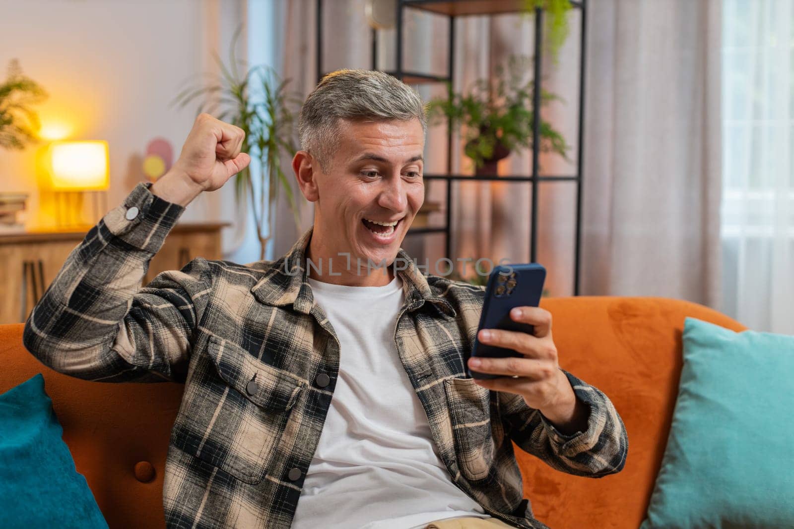 Surprised excited young man winner holding smartphone reading good news amazed by online bet bid game win. Happy guy male on sofa looking at screen overjoyed by victory success in home apartment.