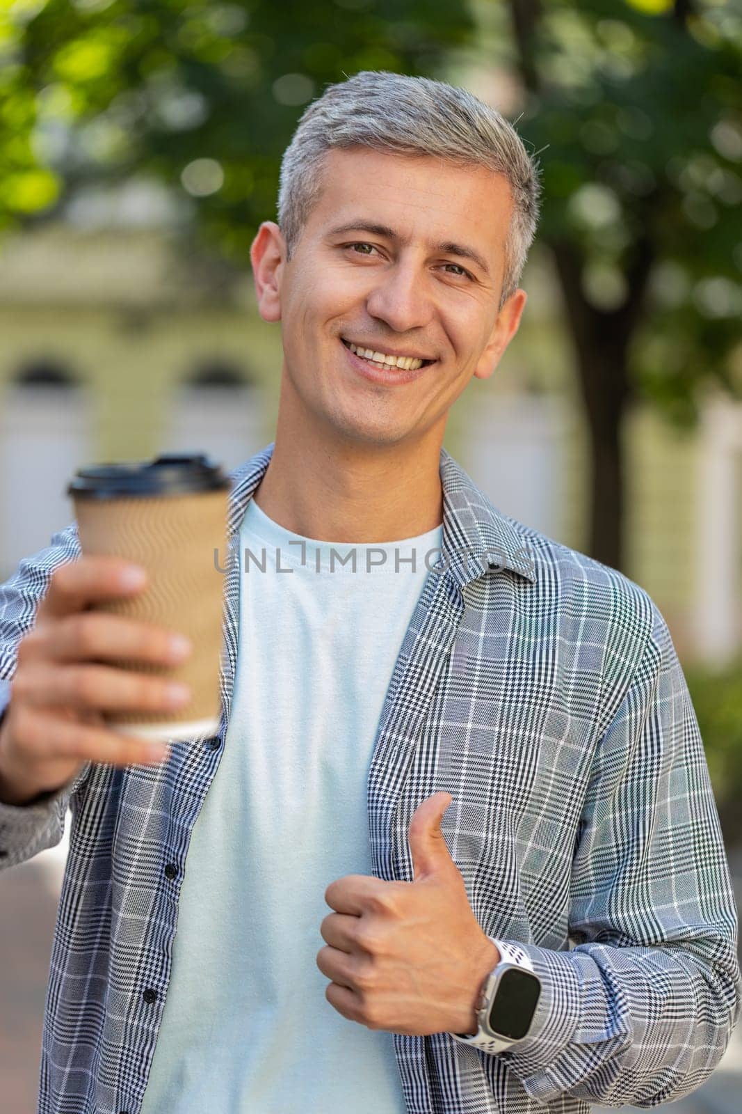 Happy Caucasian man enjoying morning coffee hot drink and smiling. Relaxing, taking a break. Mature guy standing on urban city center street, drinking coffee to go. Town lifestyles outside. Vertical