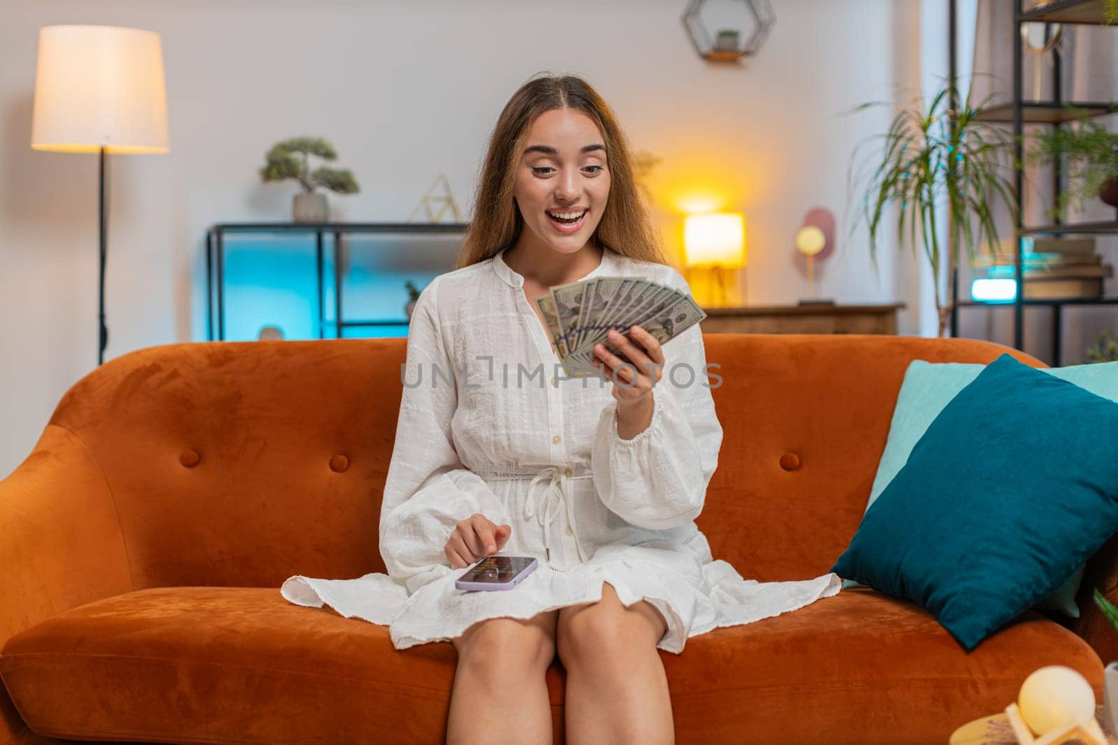 Smiling young woman counting money dollar banknotes using smartphone calculator on sofa at home. Happy adult girl calculating budget, savings, earnings and planning vacation in apartment. Lifestyle.