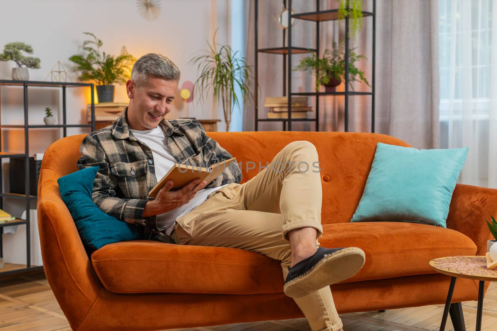 Caucasian man reading interesting book, turning pages smiling enjoying literature, taking a rest on comfortable couch. Peaceful cheerful young guy relaxing at home apartment living room on sofa