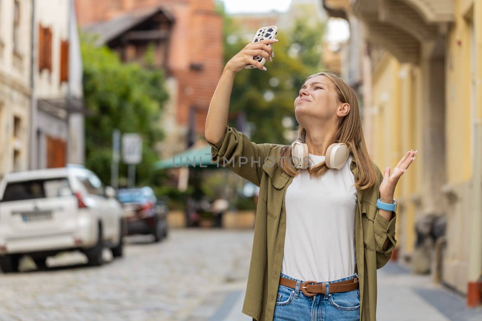 Young female tourist woman trying to catch a communication signal and Internet on mobile phone, raised the smartphone up for a better connection in city street. Frustrated Caucasian girl in town.