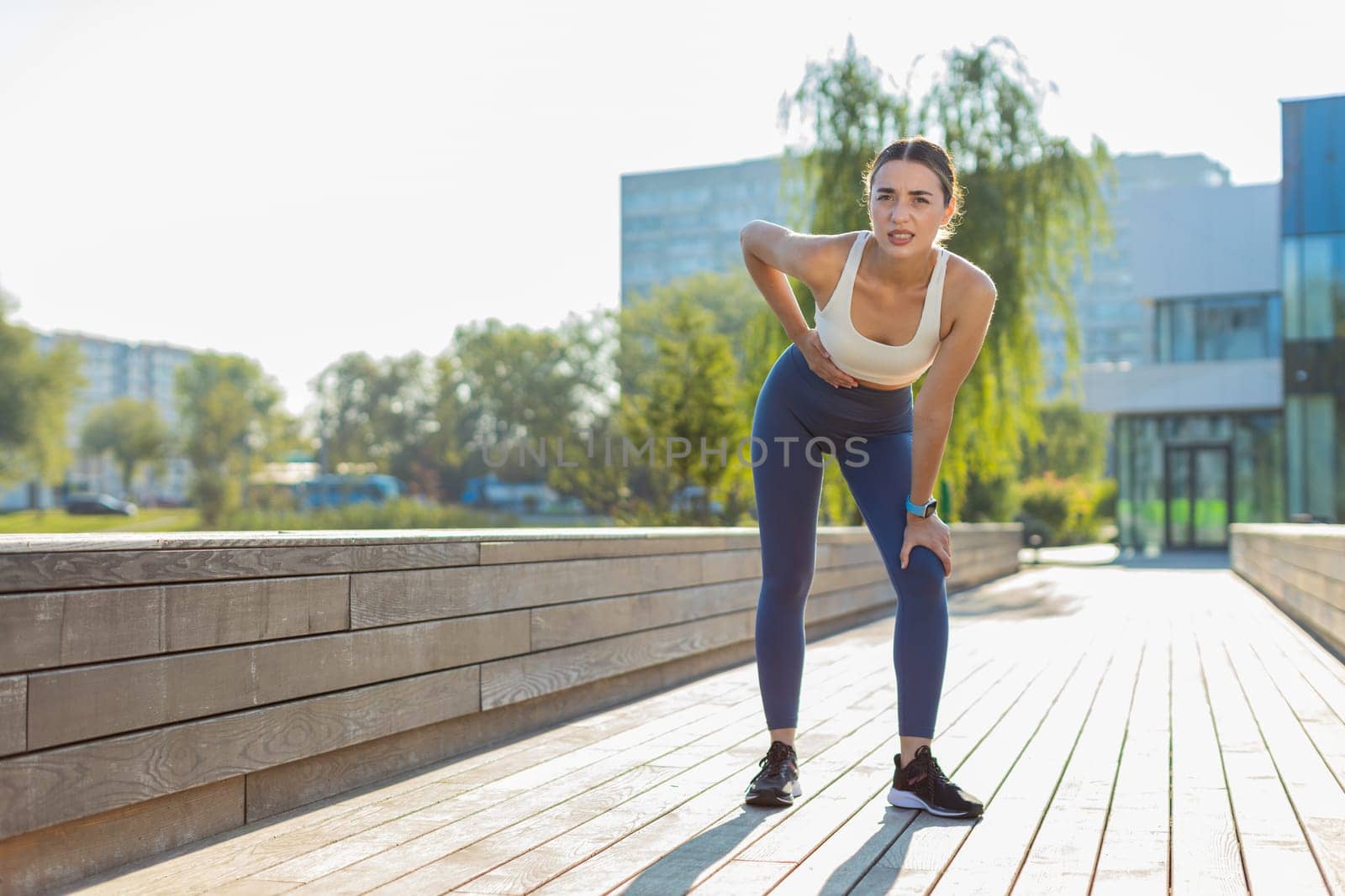 Young Caucasian runner suffering from abdominal pain during a run in urban city park on sunny day. Sad woman keeps her hand on her stomach. Girl has poisoning, constipation, spasms or menstruation.